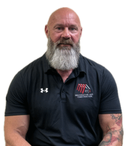 Portrait of a bald man with a full gray beard wearing a black polo shirt with a Restoration and Construction logo on the left chest.