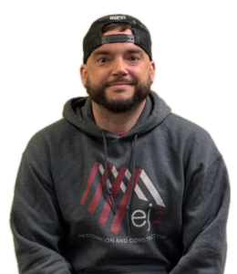 Man with beard wearing a dark gray hoodie and a black cap worn backward, facing the camera; hoodie features a red and white geometric logo and the text 'Restoration and Construction'.