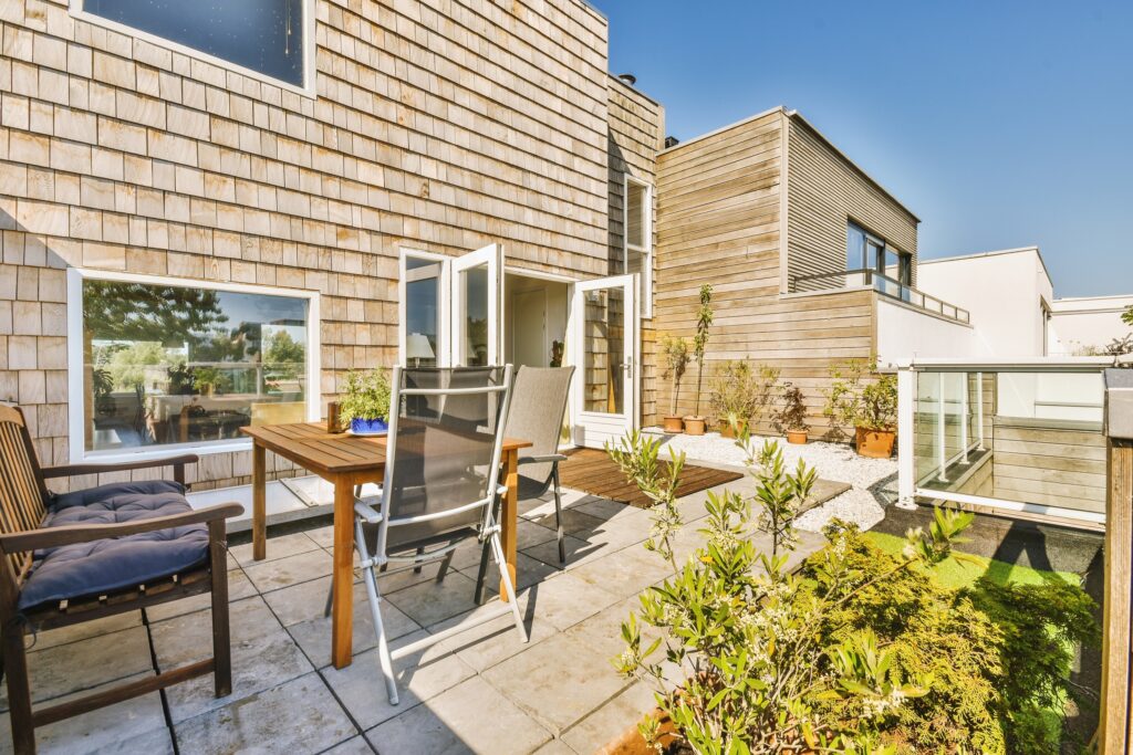 Neat patio with sitting area - EJ2 Construction Group Backyard patio of a modern wooden-clad house with an outdoor table, chairs, and potted plants under a clear blue sky.