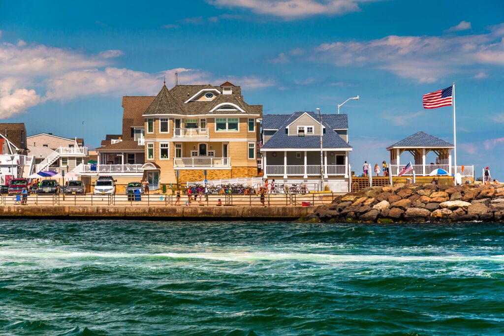 Row of beach houses along a waterfront, with a rocky breakwater and a waving American flag on a pole.