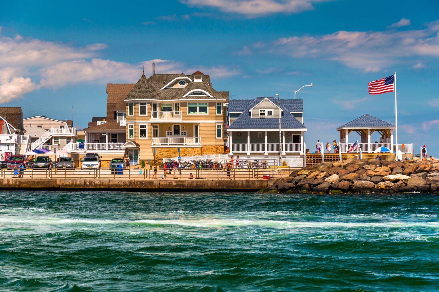 Row of beach houses along a waterfront, with a rocky breakwater and a waving American flag on a pole.