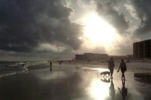 Beach scene with dark storm clouds and a bright sun breaking through, people and a dog along the wet sandy shore.