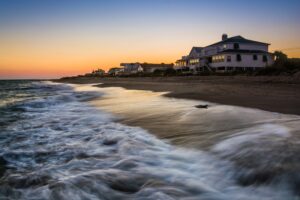 Sunset over a beach with white seaside houses on stilts along the dune, calm water and glowing orange sky.