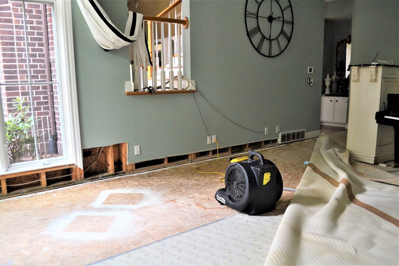 Living room under renovation with exposed subfloor, cords on the wall, and a large wall clock. A windowed door is on the left.