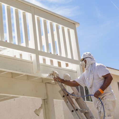 House Painter Wearing Facial Protection Spray Painting A Deck of A Home. House Painter Wearing Facial Protection Spray Painting A Deck of A Home.